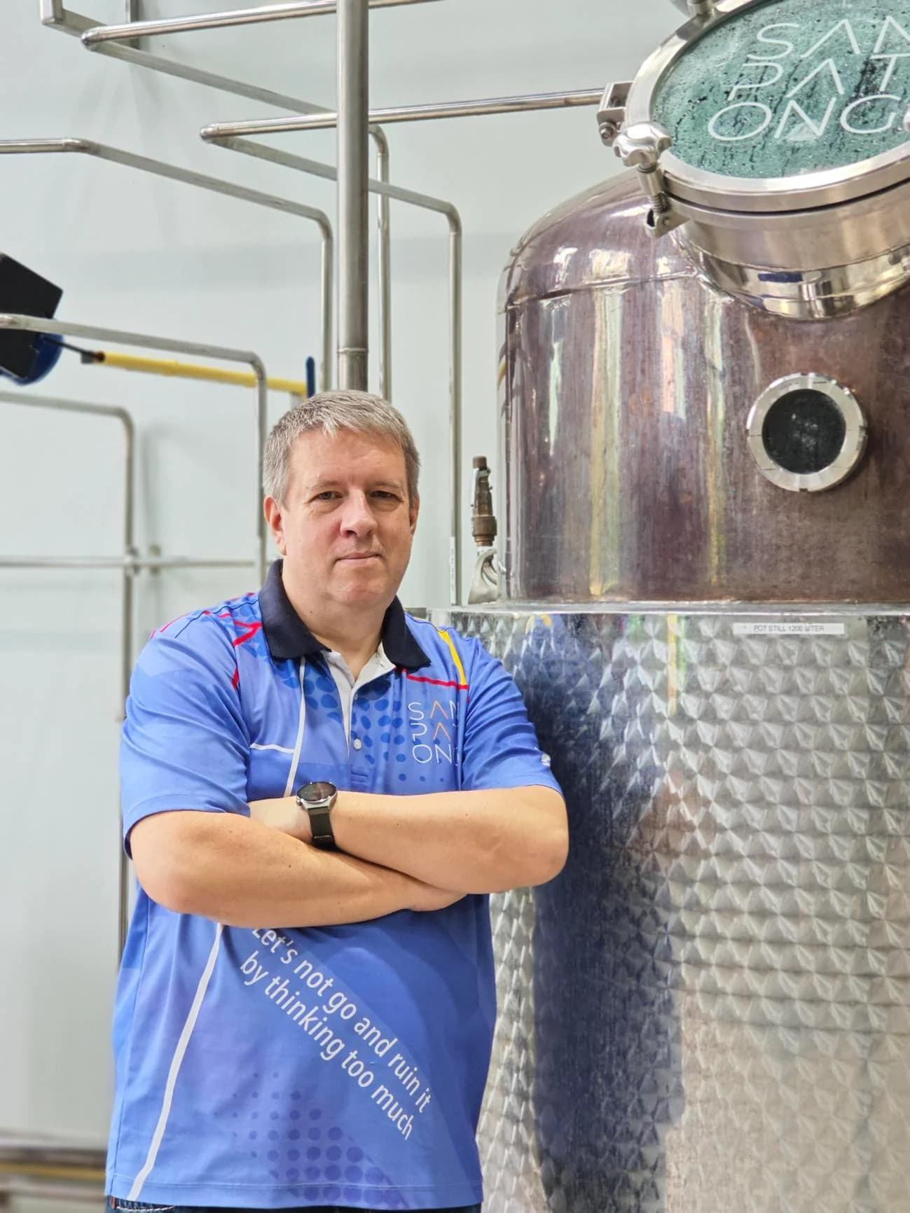 Man in blue uniform standing with arms crossed beside a large stainless steel distillation tank in an industrial facility