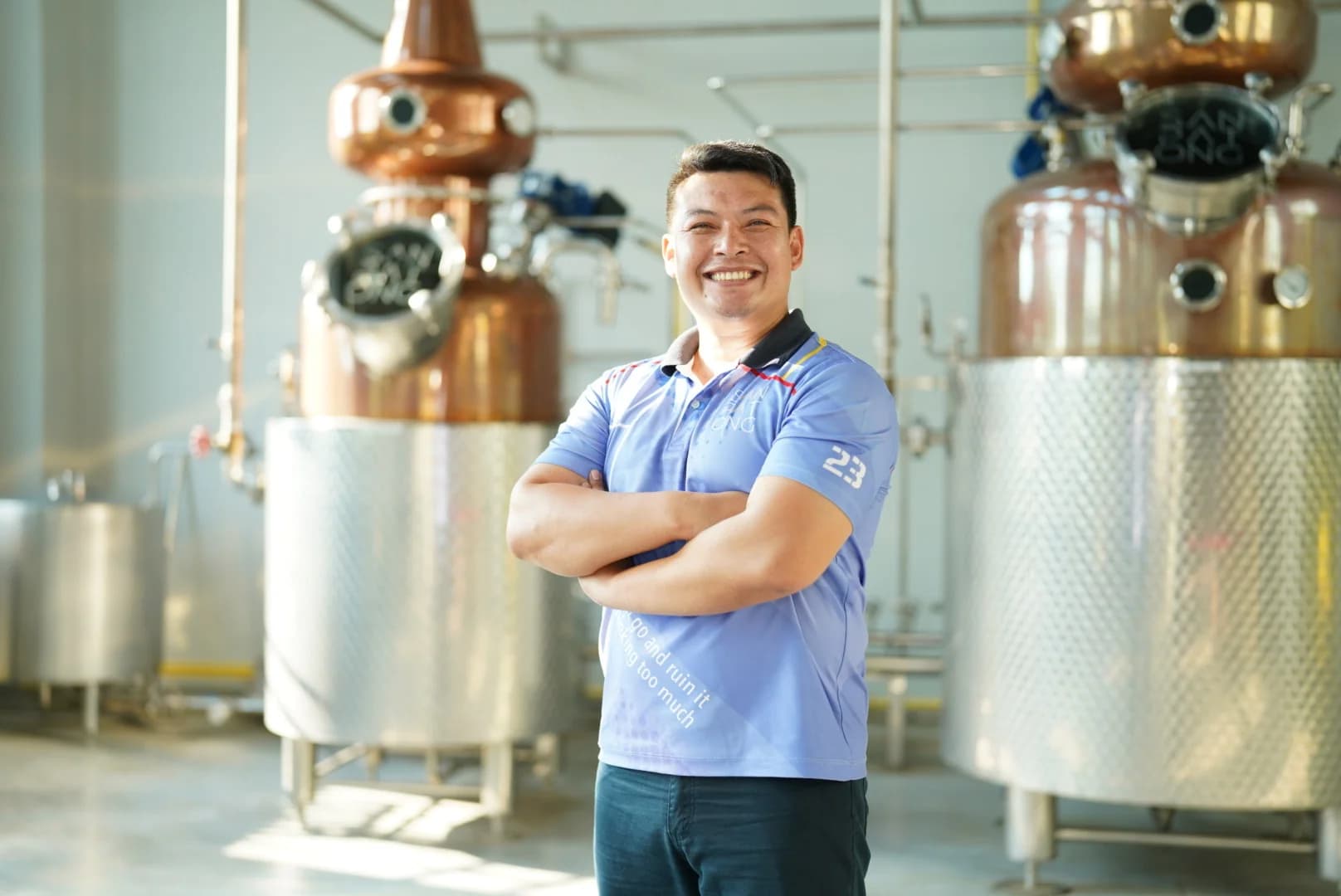 Smiling distillery director with arms crossed standing in front of stainless steel and copper brewing tanks in a modern production facility.