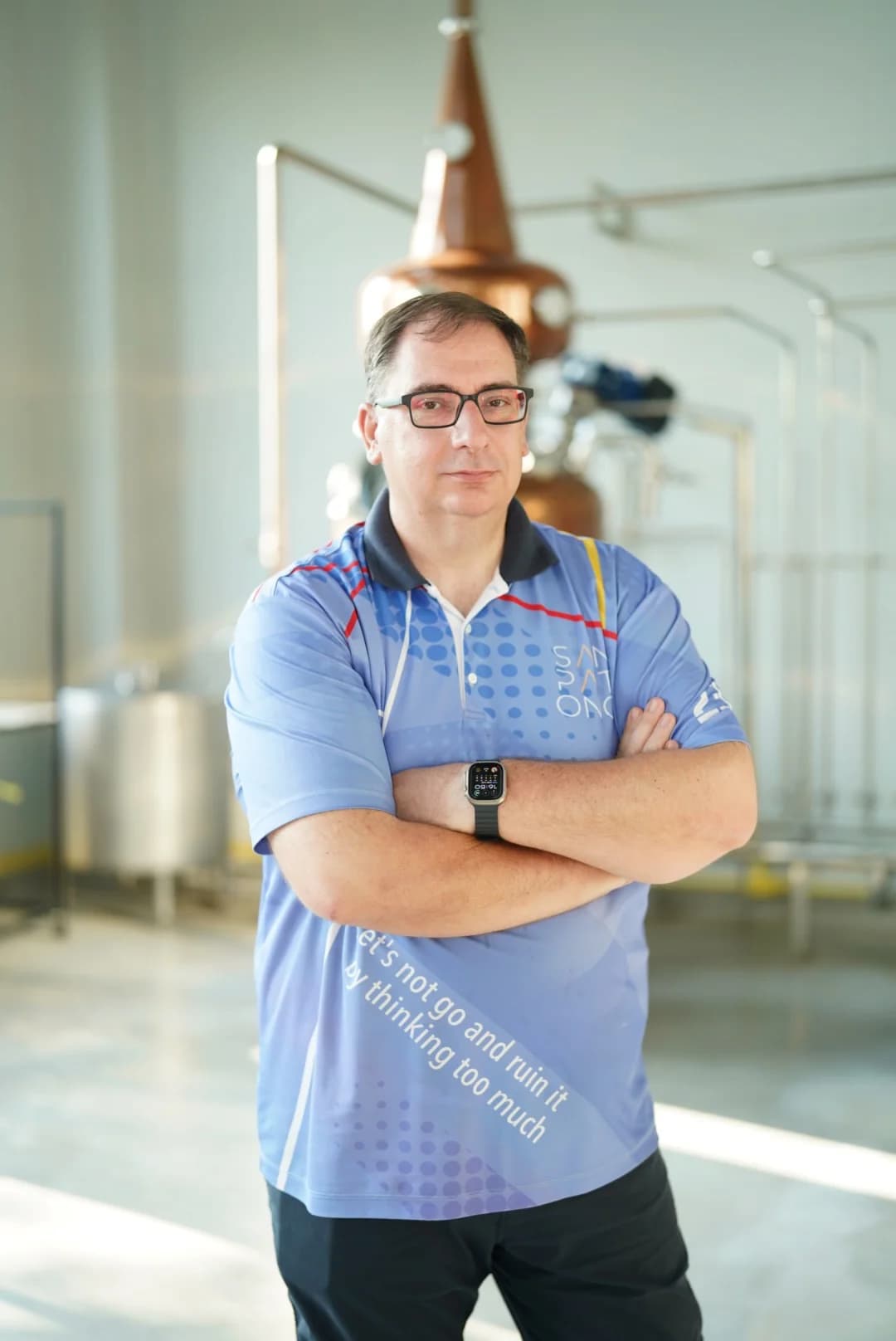 Stefan Rakoczi with glasses in blue polo shirt standing arms crossed in a distillery with copper still and stainless steel tanks in background