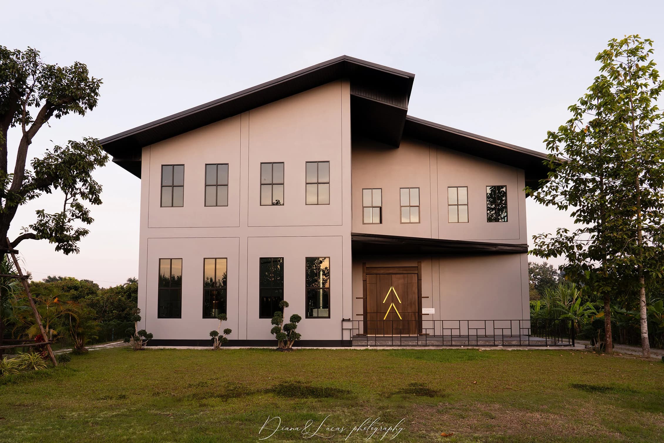Modern two-story distillery with white facade, large windows, and a dark sloped roof, set on a grassy lawn with trees at sunset.
